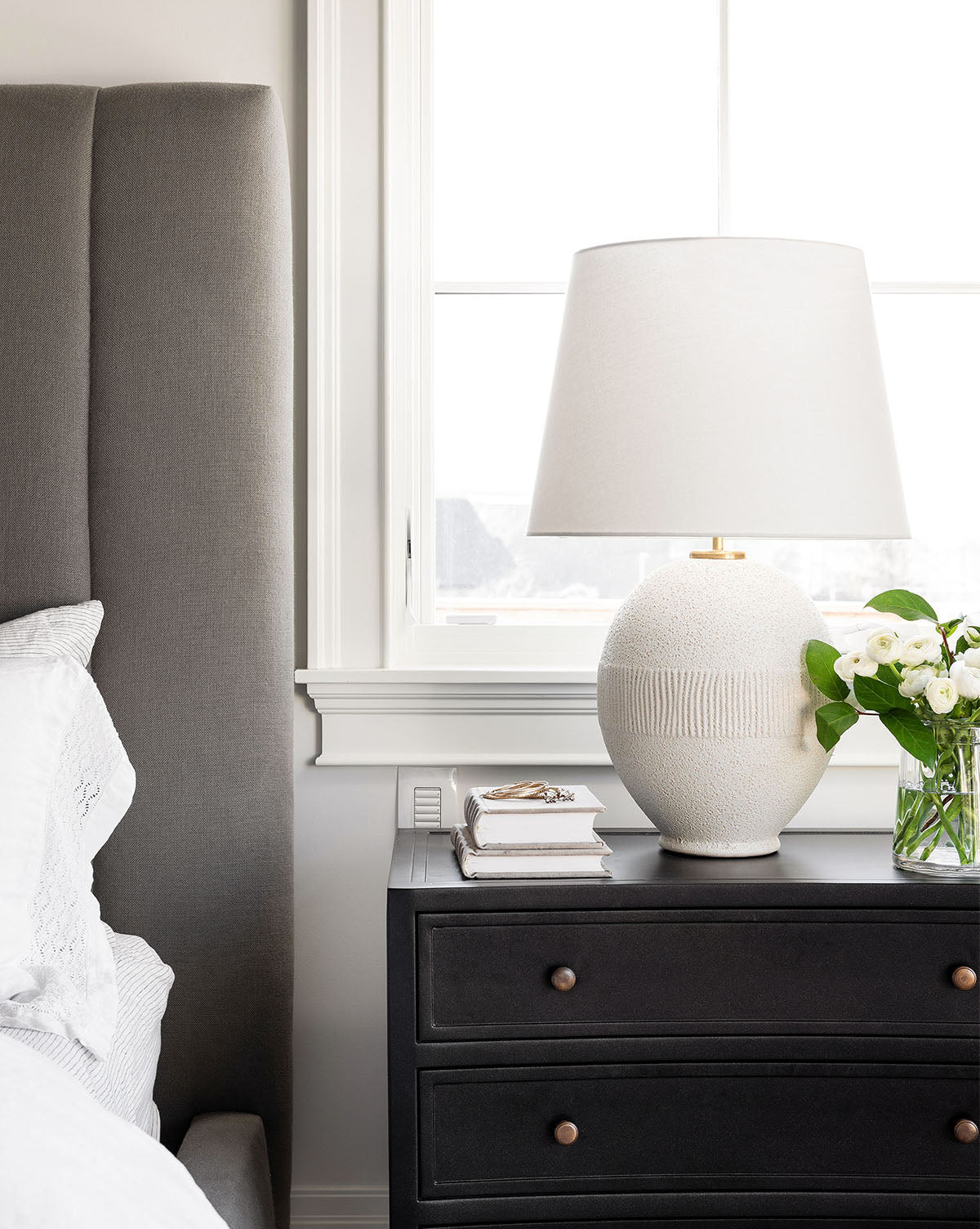 A bedside table with a natural texture features the Toulon Table Lamp by Visual Comfort & Co. Signature with a linen shade, alongside a vase of white flowers, books, and glasses next to a gray upholstered bed in a modern bedroom. Vendixs