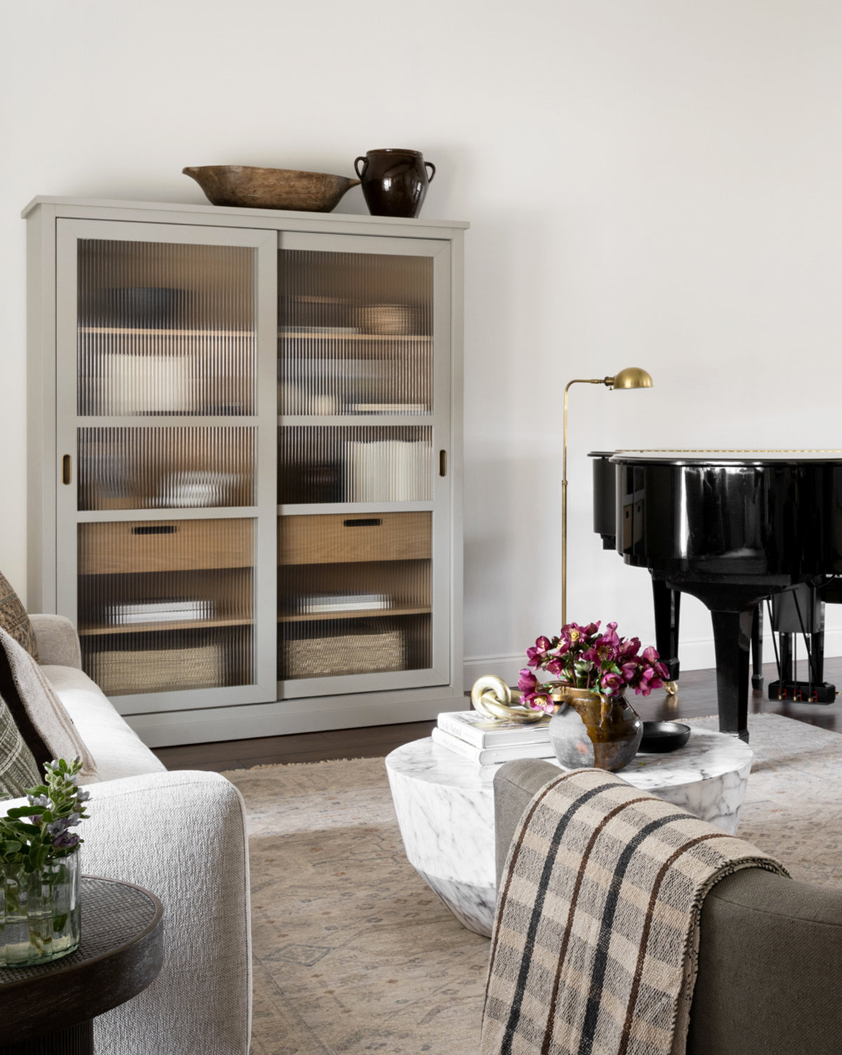 A cozy living room with a white cabinet of baskets, a marble coffee table with books and flowers, a beige sofa with a plaid throw, the Old Pharmacy Floor Lamp for adjustable height lighting, and a black grand piano against a white wall, by Vendixs