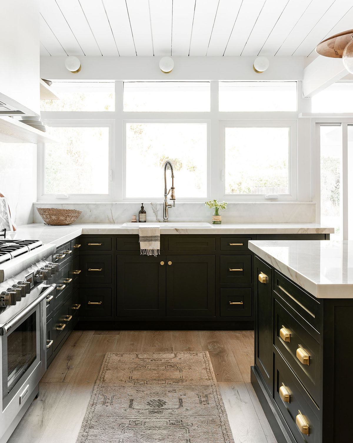 Bright modern kitchen with black cabinets, gold hardware, white countertops, stainless steel appliances, a farmhouse sink, large windows, and an Ellington Fawn Hand-Tufted Wool Rug on light wood floors, by Vendixs