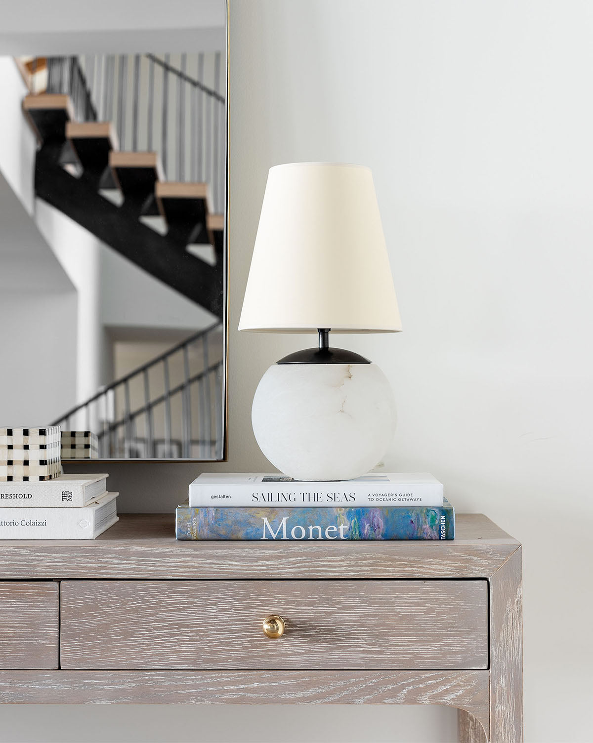 A Terri Round Table Lamp with a white shade and alabaster base sits on stacked books, including one titled Monet, on a wooden console table. Behind it, a large mirror reflects stairs with black railings, by Vendixs