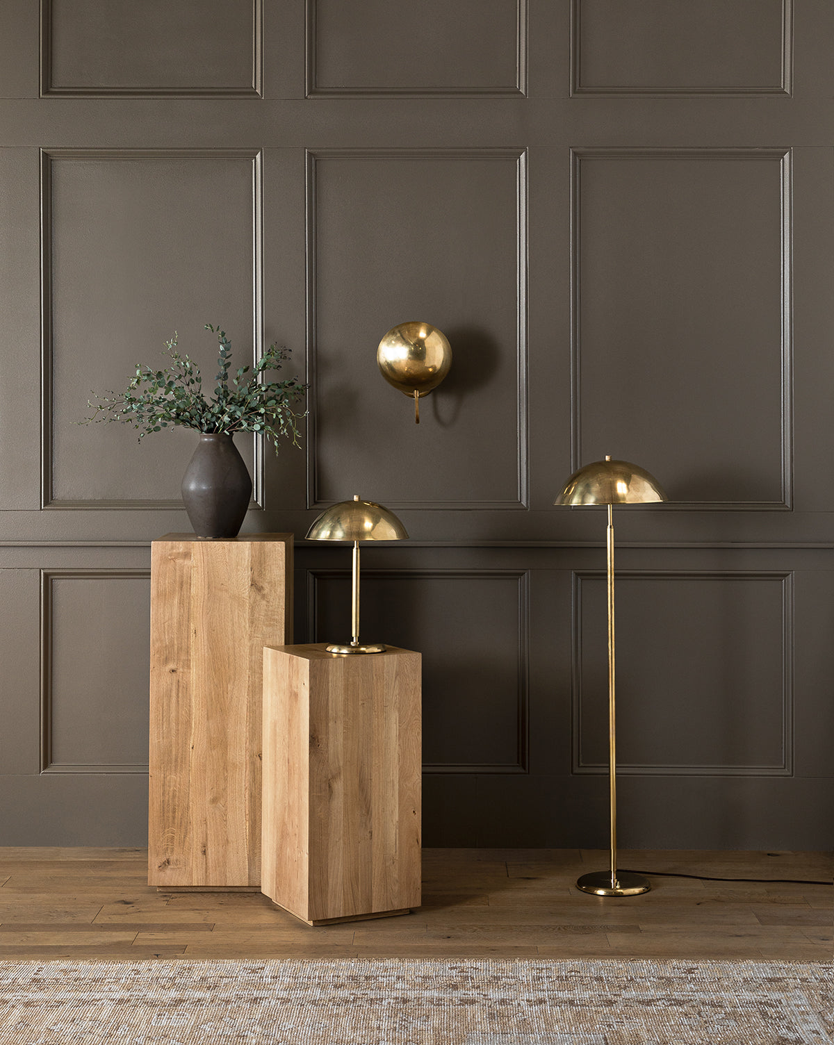 A modern interior with a dark paneled wall, two wooden pedestals, a black vase with greenery, and three gold dome-shaped lamps—including the elegant Aldorno Floor Lamp—creates a sophisticated, stylish atmosphere, by Vendixs