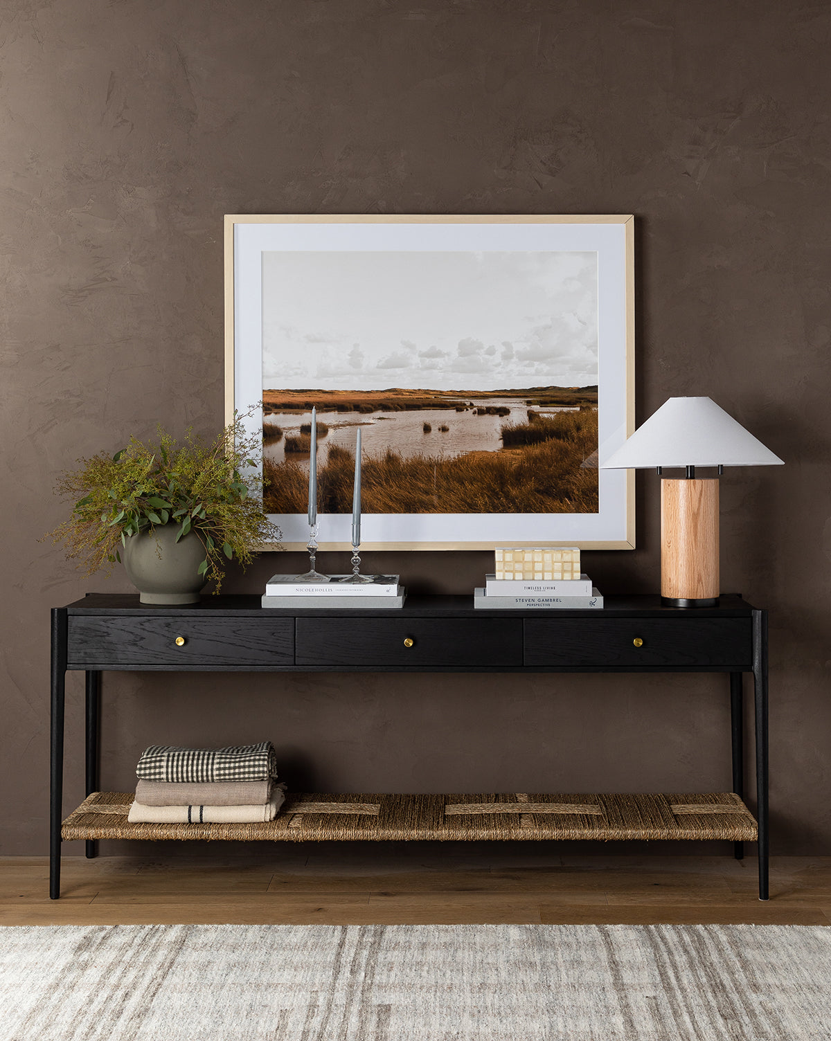 The Dana Console in black with brass hardware holds a lamp, vase with plant, candlesticks, books, and decor. A framed landscape photo hangs above. Blankets are folded on the woven shelf below—a perfect entryway accent, by Vendixs