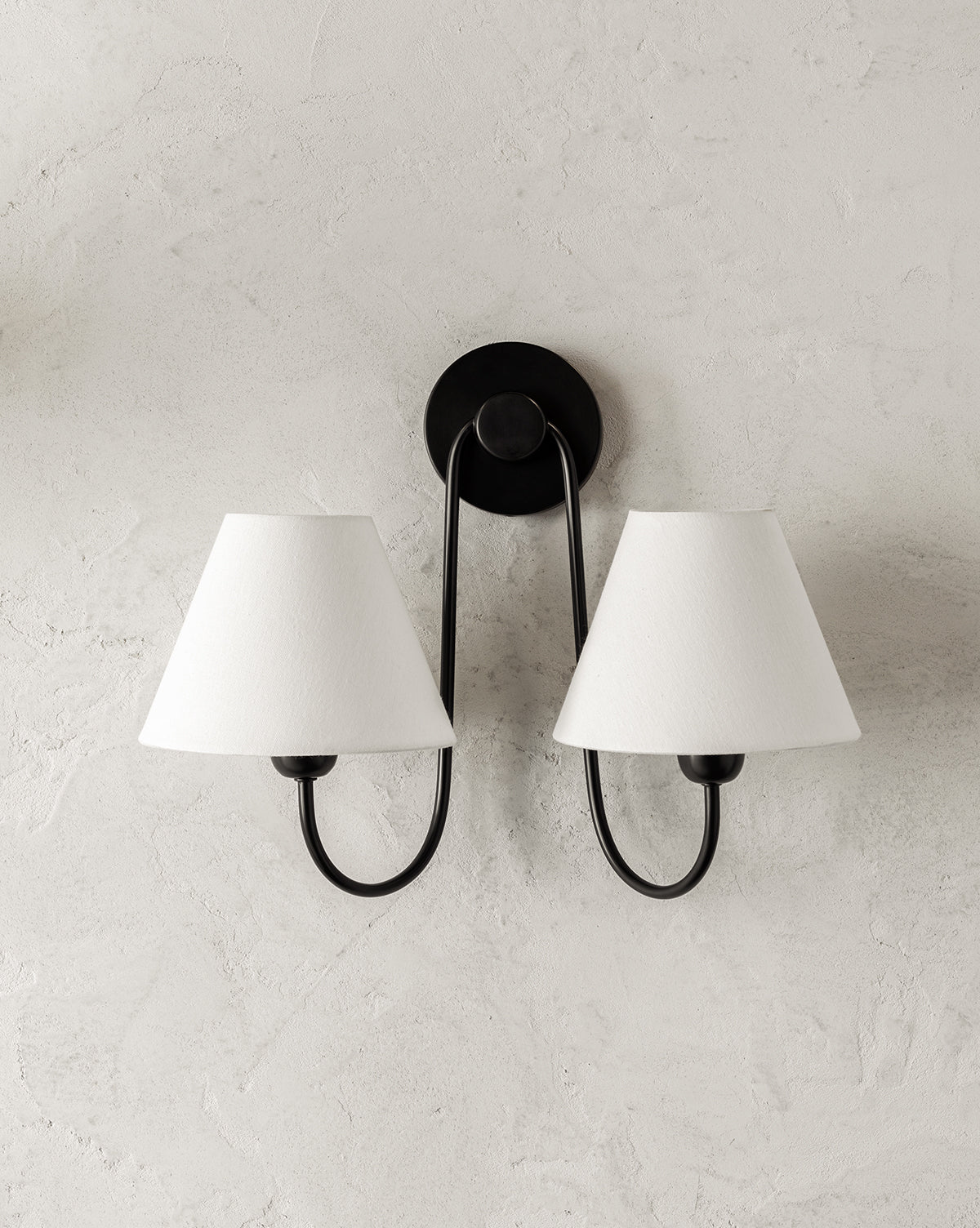The Wainwright Double Swoop Sconce by Makerspalm features two black metal arms and white linen shades, elegantly mounted on a light textured wall. Vendixs