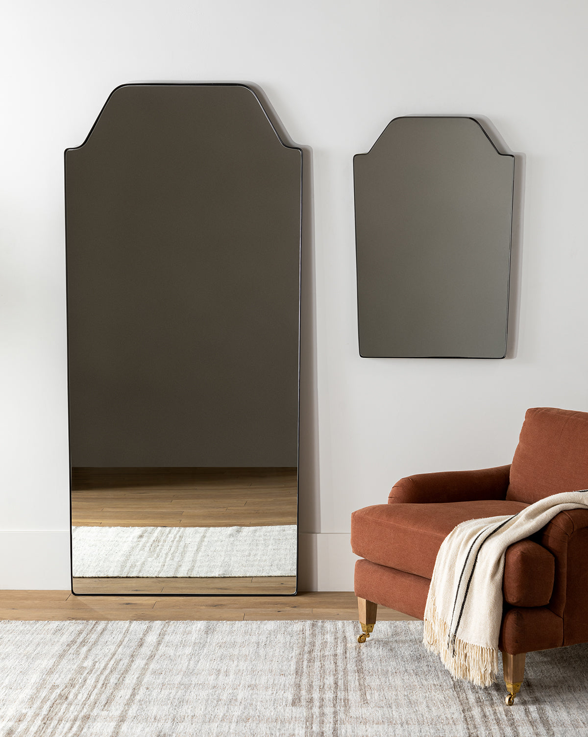 A modern room features the tall matte black Giles Floor Mirror with a geometric top, paired with a matching wall mirror, beside a rust armchair draped in a light throw on a neutral rug over wood flooring, by Vendixs