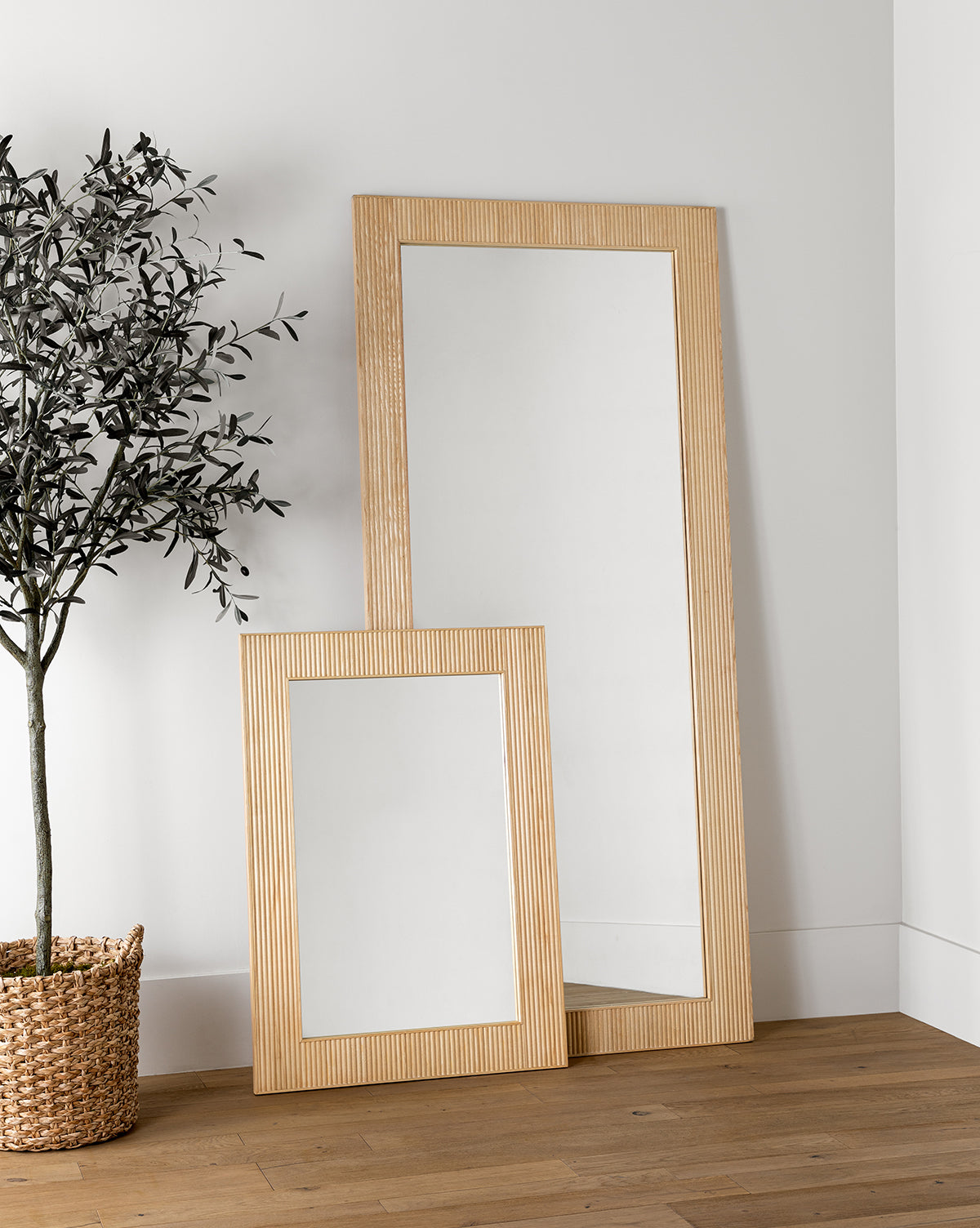 Two Rowan Wall Mirrors with light ribbed ash wood frames lean against a white wall on a wooden floor. A potted plant in a woven basket accents these modern Rowan Wall Mirrors, by McGee & Co.
