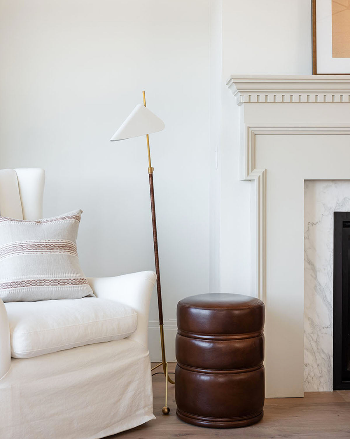 A cozy white armchair with a patterned pillow sits beside the Moresby Floor Lamp, offering stylish lighting, and a round brown leather ottoman is placed near a white fireplace with a marble surround, by Vendixs
