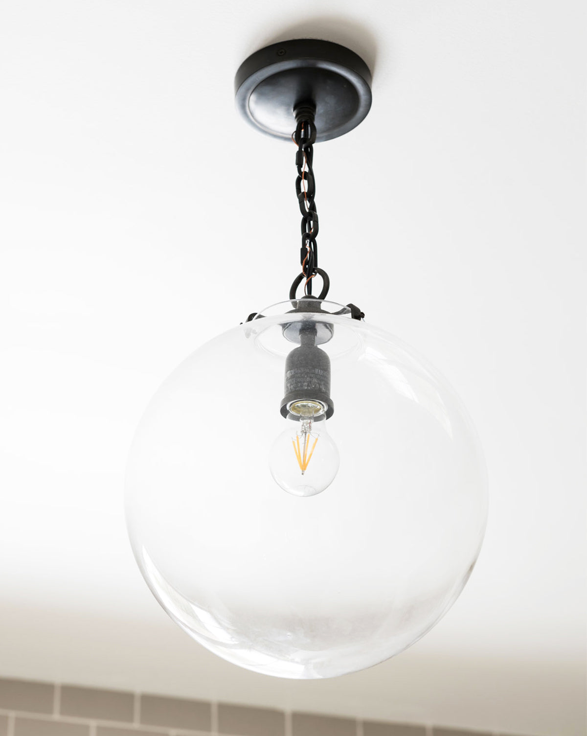 The Katie Globe Pendant by Visual Comfort & Co. Signature features a round glass globe suspended from a short black chain on a white ceiling, highlighting an exposed bulb inside, by Vendixs