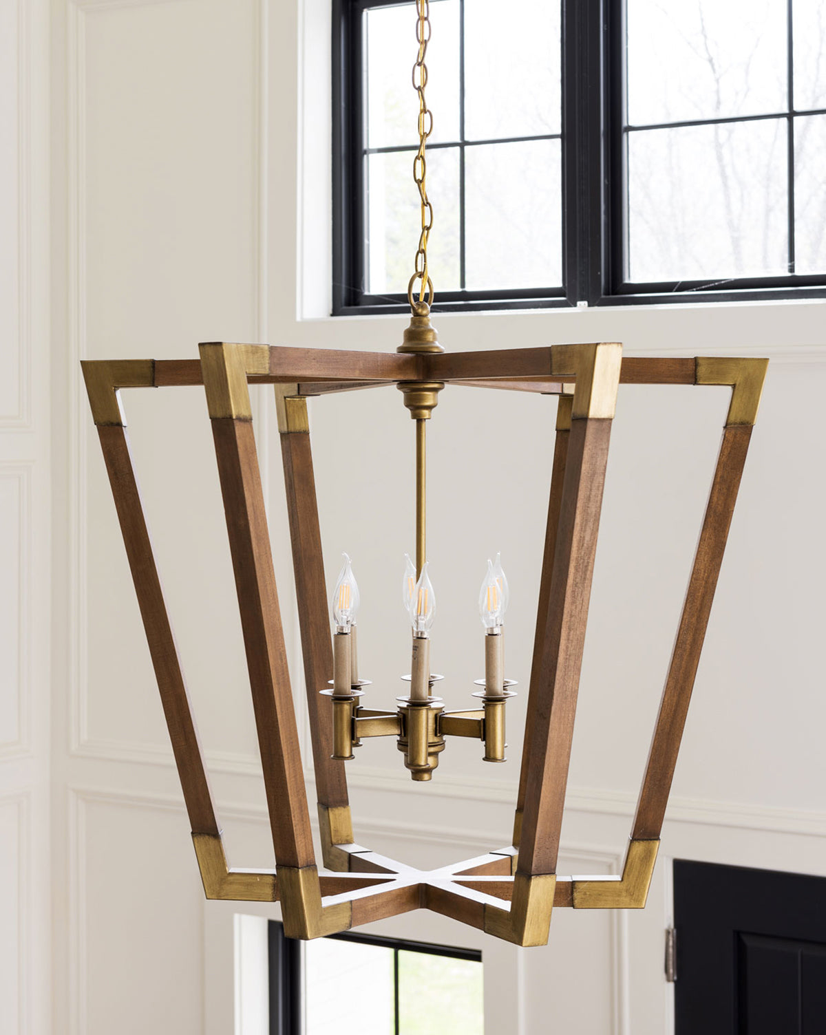 The Bastian Chandelier by Currey & Co. features a geometric chestnut and brass frame with four candle-style bulbs, adding modern elegance suspended near large black-framed windows. Vendixs