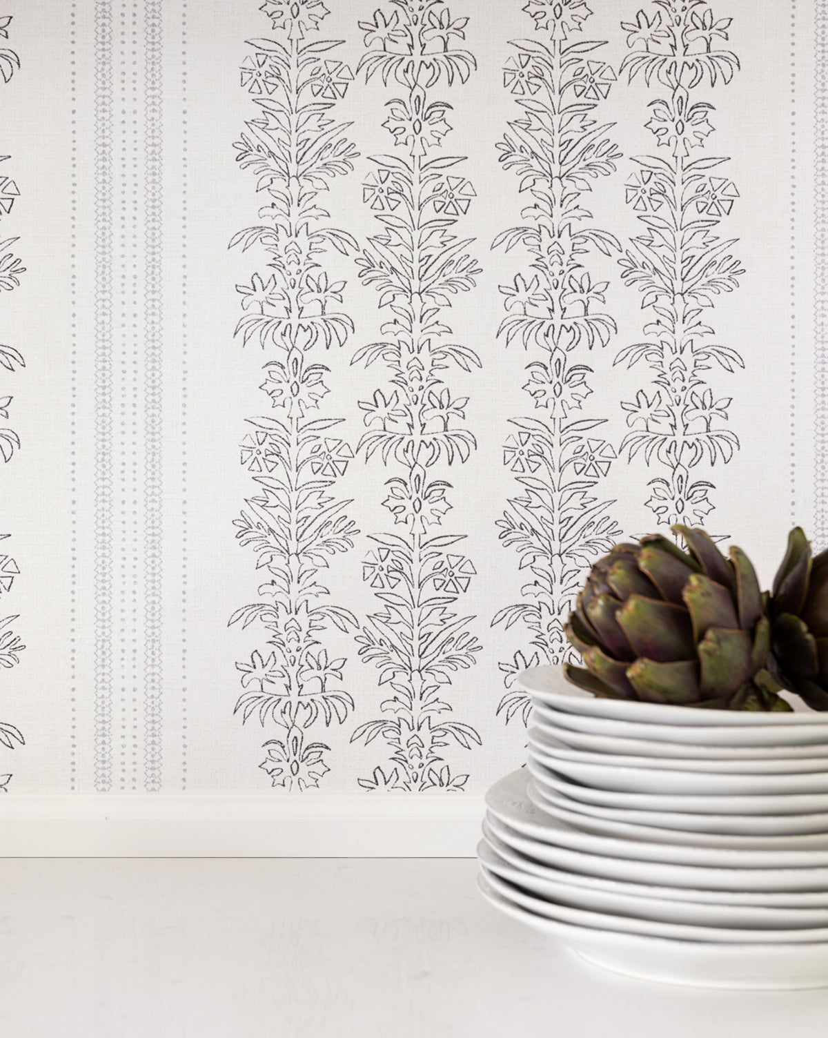 A stack of white plates sits on a white surface, topped with an artichoke, in front of a wall adorned with elegant floral motifs and delicate York Wallcoverings Demi Floral Stripe Wallpaper, Vendixs