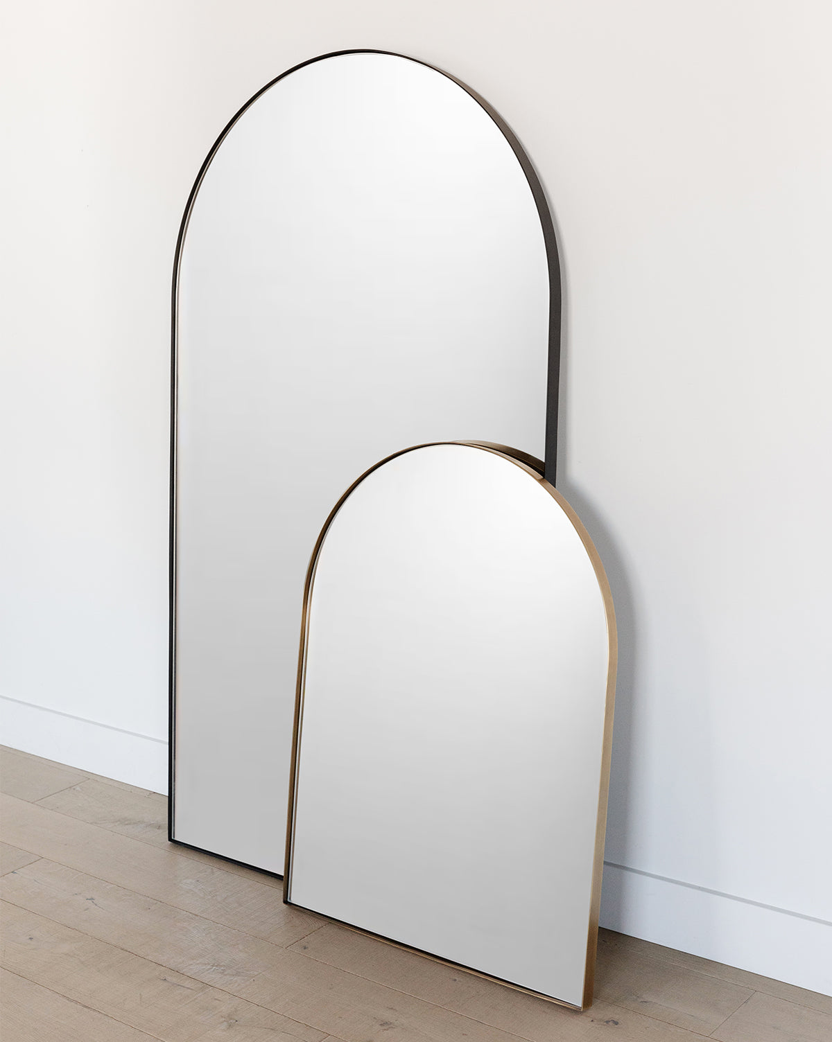 Two Wylie Arched Mirrors with slim metallic frames and shatterproof glass lean against a white wall on a light wooden floor. The taller entryway mirror is partially overlapped by the shorter one, creating a modern layered look, by Vendixs