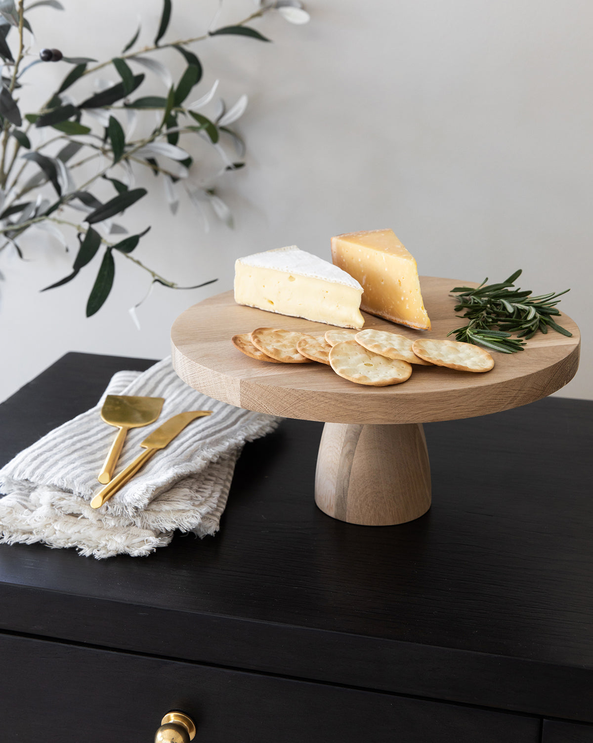 A Wooden Cake Stand displays two cheese wedges, round crackers, and rosemary sprigs. Gold utensils on a folded napkin sit on a dark side table, with leafy branches in the background for stylish home decor by Vendixs