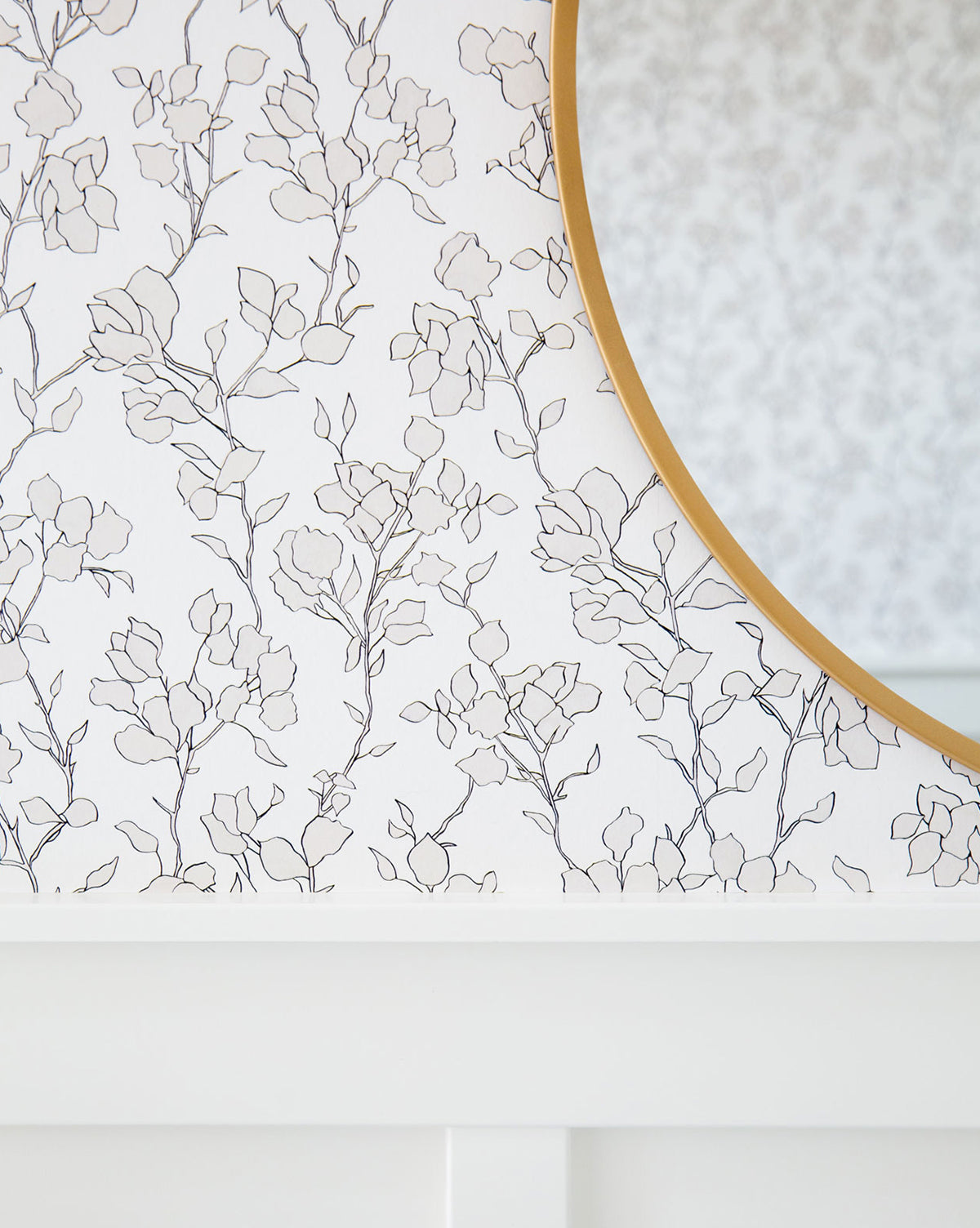 A round mirror with a gold frame hangs on a wall adorned in Blair Sketched Floral Wallpaper by York Wallcoverings, featuring delicate black lines on white. Elegant white wood molding is visible at the bottom of the image. The overall aesthetic evokes the timeless style of Vendixs
