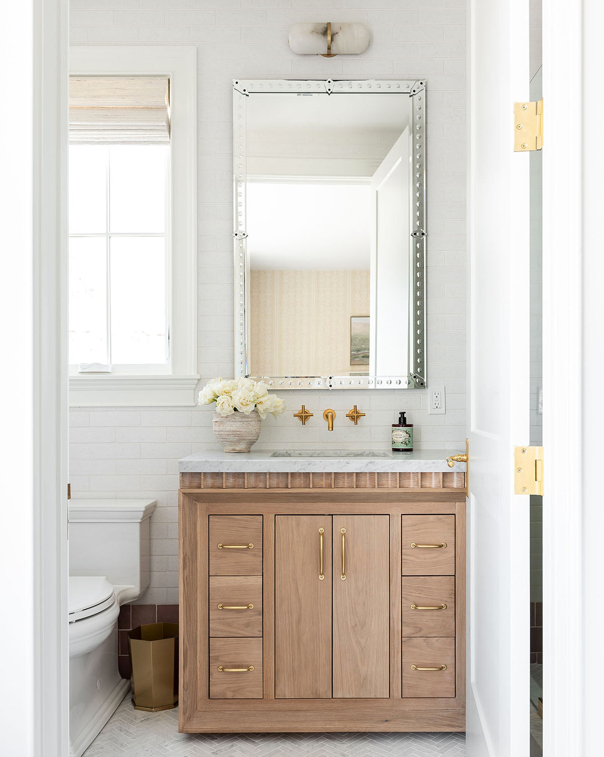 A bright bathroom showcases a light wood vanity with gold hardware, white countertops topped with the Truman Mirror, a wall-mounted gold faucet, a white flower vase, and a toilet beside the window, by Vendixs